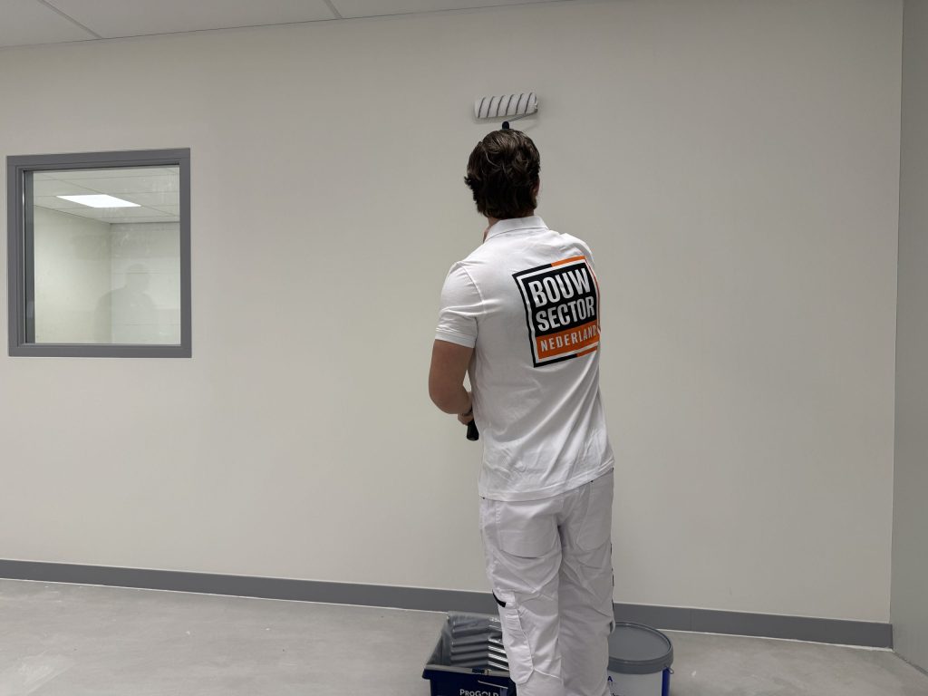 Schilder in witte werkkleding zet de eerste baan verf op een lichte wand in een lege ruimte