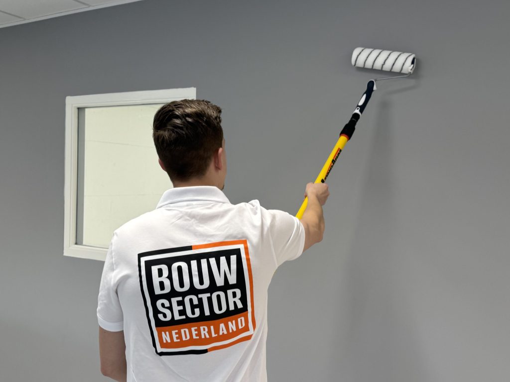 Schilder met het logo van Bouwsector Nederland op zijn rug rolt een grijze muur naast een raam met wit kozijn