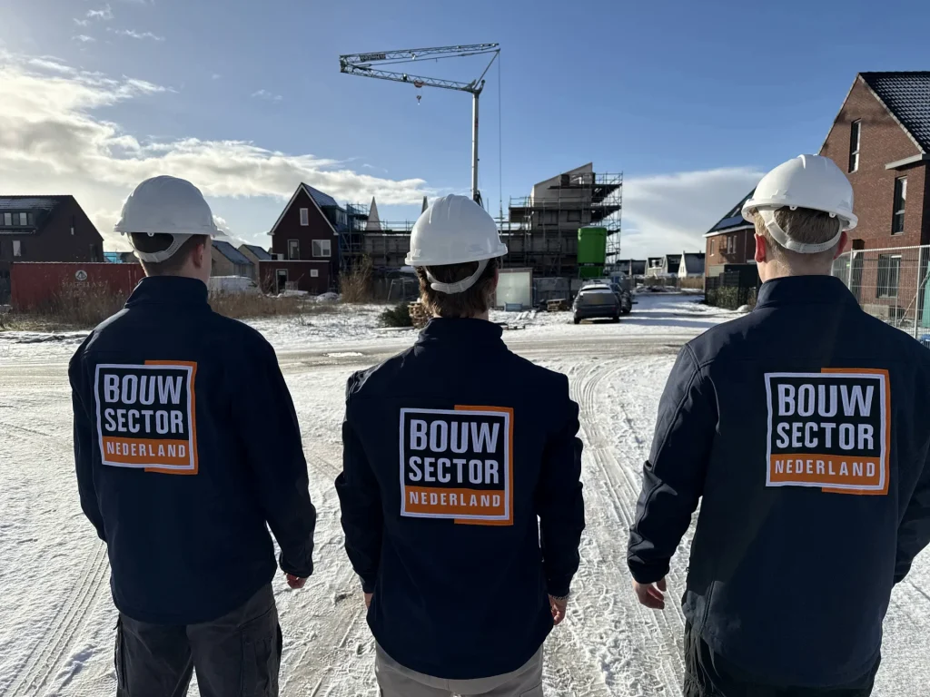 Drie vakmensen van Bouwsector Nederland in donkerblauwe jas met logo kijken naar een nieuwbouwwijk