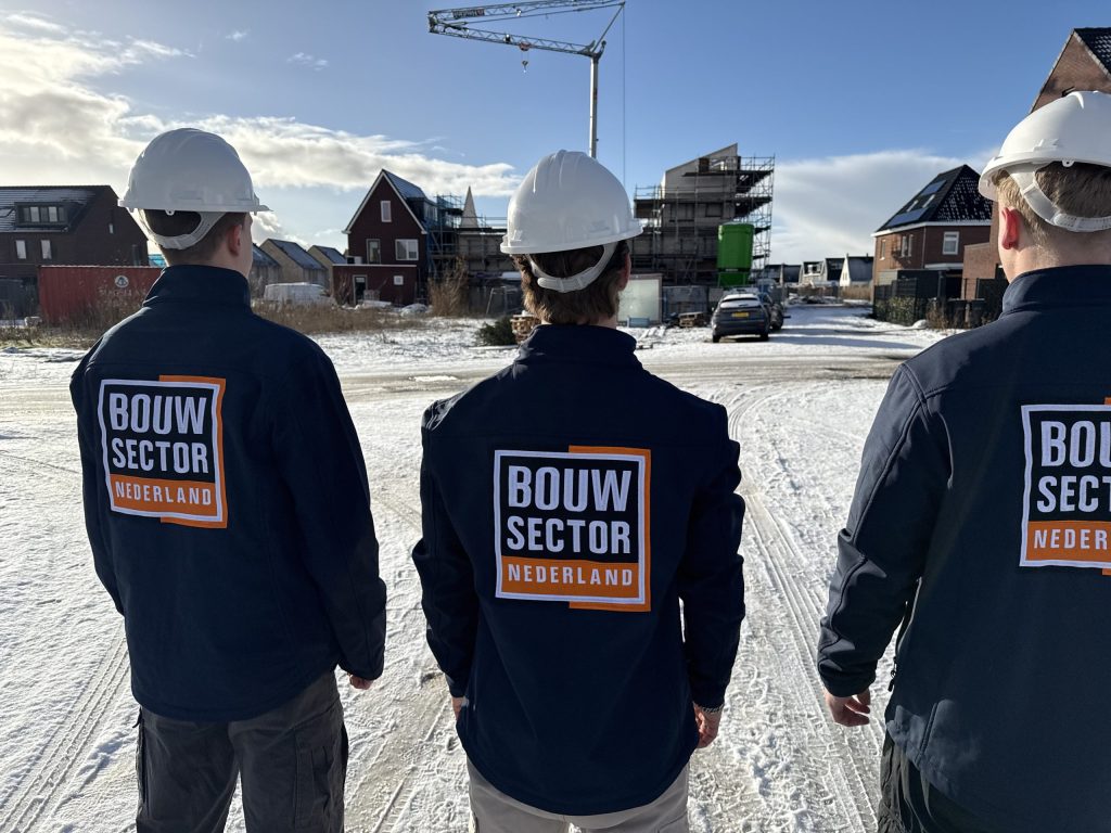 Drie vakmensen van Bouwsector Nederland met witte helm lopen door een besneeuwde straat met nieuwe bakstenen huizen