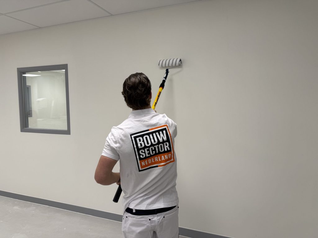 Schilder van Bouwsector Nederland rolt muurverf op een lichte wand naast een binnenraam